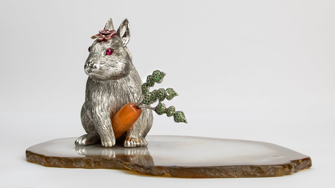 Front view silver sculpture depicting a winter rabbit holding a carved carnelian and tsavorite carrot in its paws, ruby eyes, fancy sapphire flowers, on a slice of agate