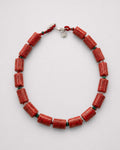 A beaded necklace made of cylindrical red coral-like beads separated by small dark green spacer beads, with a silver clasp and red cord, shown on a light gray background.