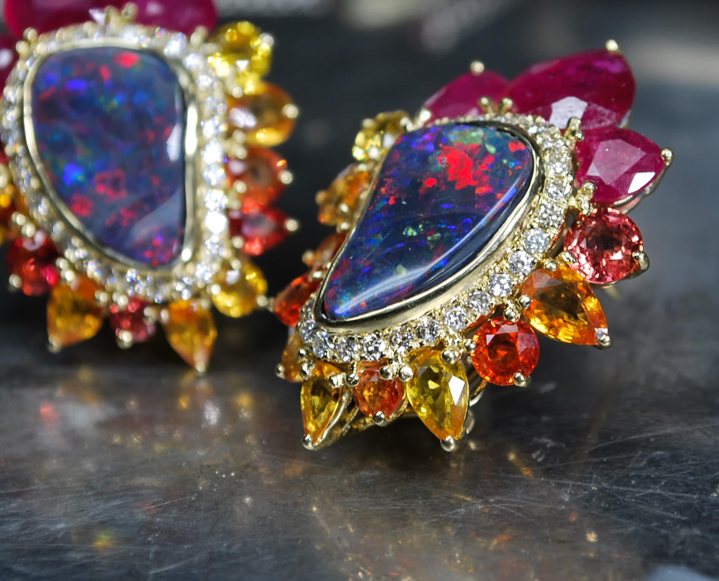A pair of ornate gold earrings featuring large teardrop-shaped iridescent opal gemstones, surrounded by round and marquise-cut pink, orange, yellow, and clear accent stones on a dark reflective surface.