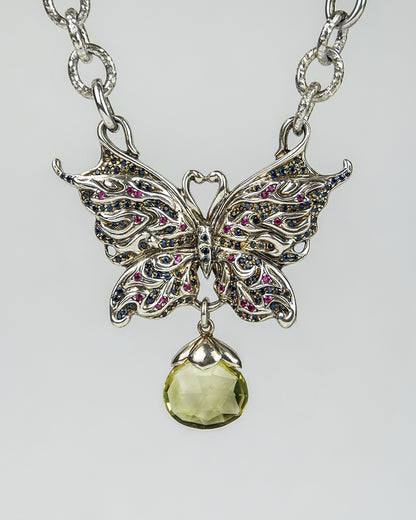 close-up of a silver necklace, featuring blue sapphires and rubies set in a butterfly-shaped pendant, complemented with a briolette citrine dropping from the centre, hanging from a textured silver chain