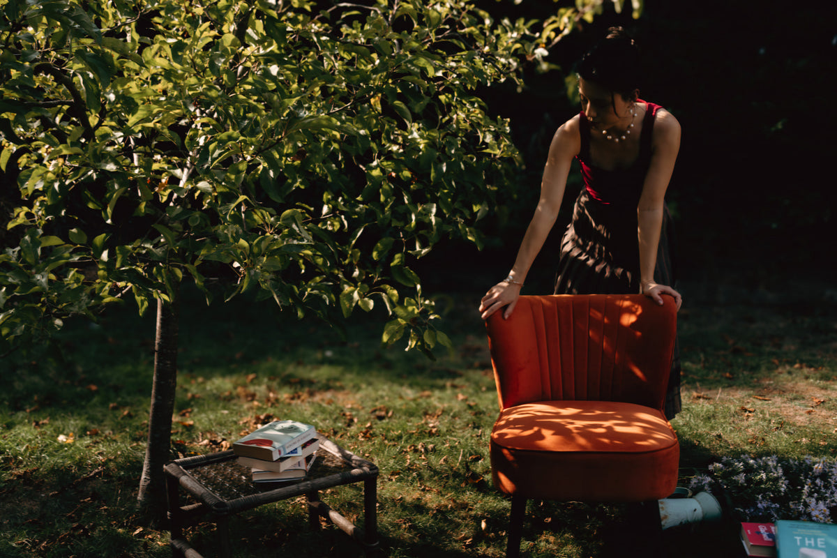 A woman in a dark dress stands beside an orange upholstered armchair in an outdoor garden setting with a tree and sunlight filtering through the leaves.
