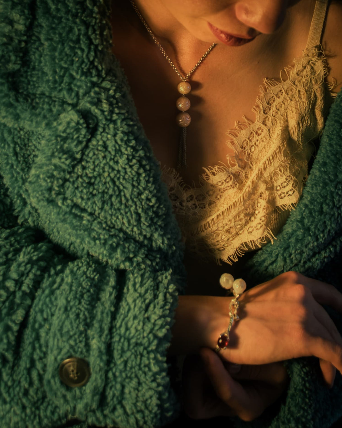 Person wearing silver necklace with pearls and bangle from TVRRINI, complemented by a chunky green jacket and cream lace camisole.