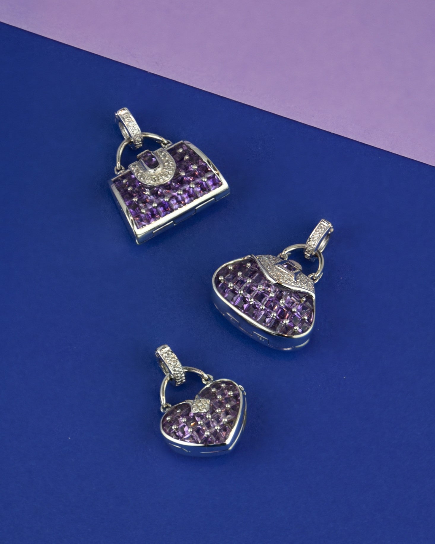 Three unique TVRRINI 18K white gold with amethyst and diamond locket pendants on blue paper next to lilac paper