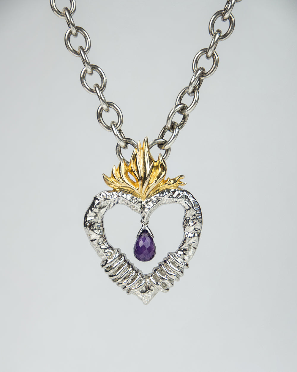 front side of a silver pendant, featuring a briolette-cut amethyst hanging in the middle of a heart with a flame at the top plated with yellow gold