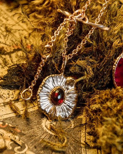 angled view of a pendant featuring garnet oval cabochon, resting in an indoor setting with grass and printed paper