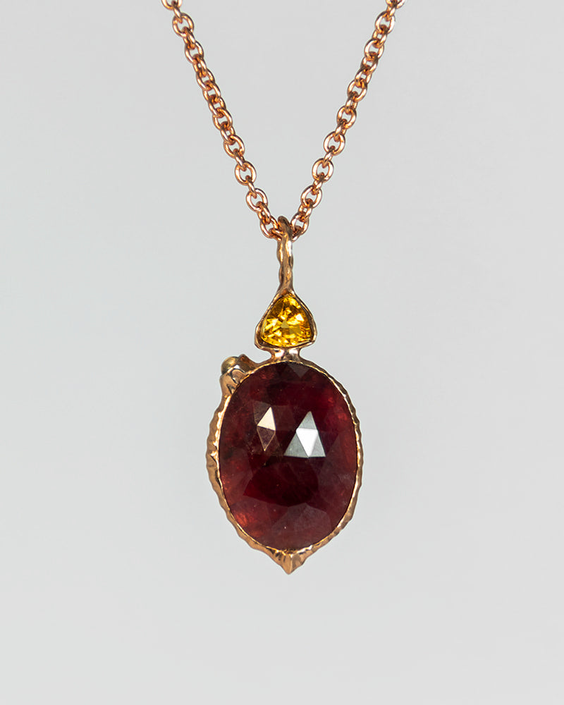 Rose gold-plated pendant necklace featuring a large oval faceted dark red gemstone and a small trillion-cut yellow gemstone.