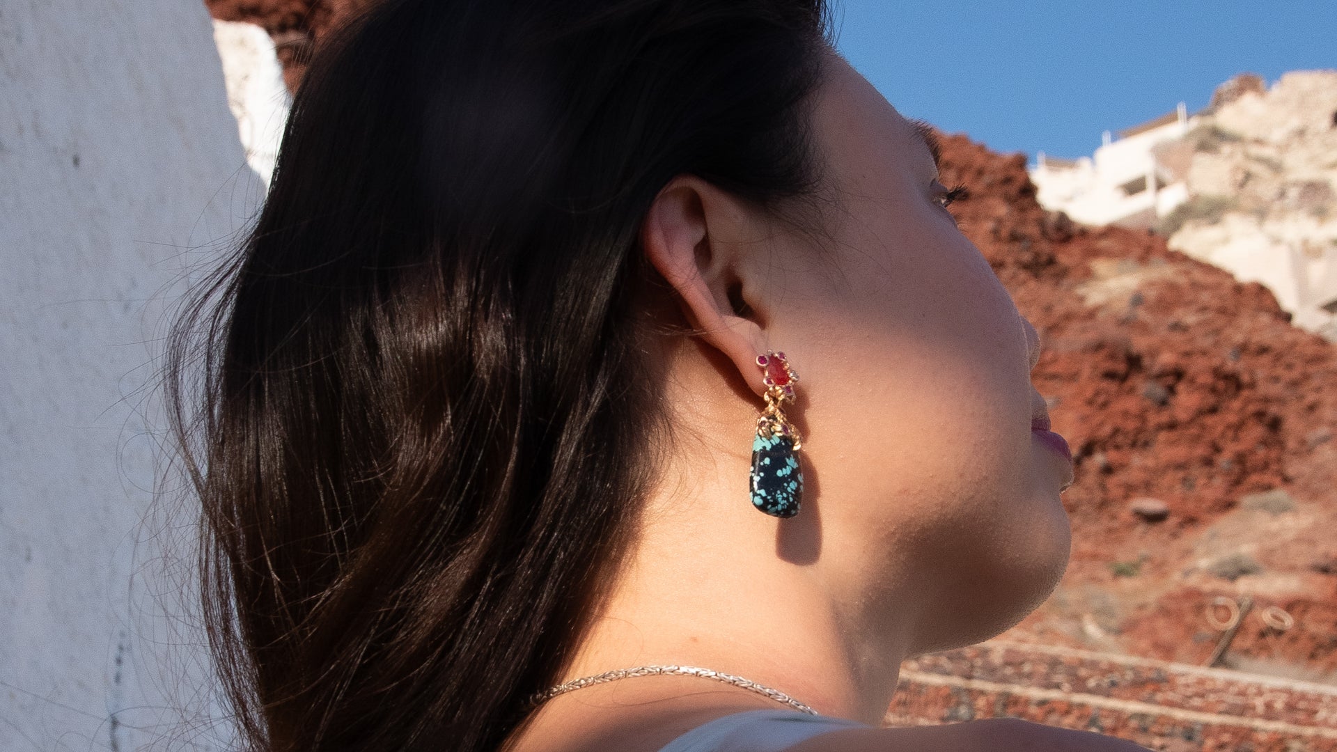 Person wearing unique Lava Earrings pair, the red mountains of Santorini behind