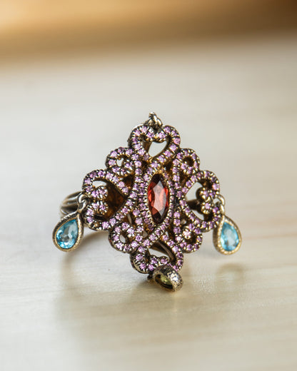 close-up of a silver ring, featuring rhodolite pavé set, with dangling pear-cut blue topaz and peridot 