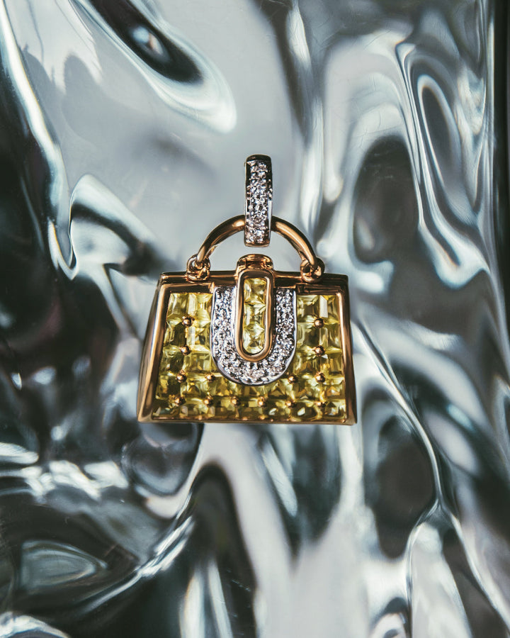 close-up of a handbag-shaped 18K gold locket featuring peridots and diamonds with a polished finish, against a resin sculpture