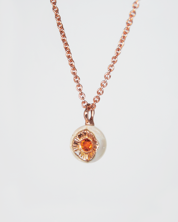 angled view of a pendant with a round freshwater pearl set with a round faceted orange sapphire held together by a sculptural gold-plated silver design