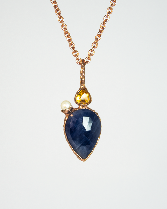 front view of a pendant featuring a teardrop faceted blue sapphire, trillion-cut citrine, and white freshwater pearl nestled in an abstract setting made from gold-plated sterling silver